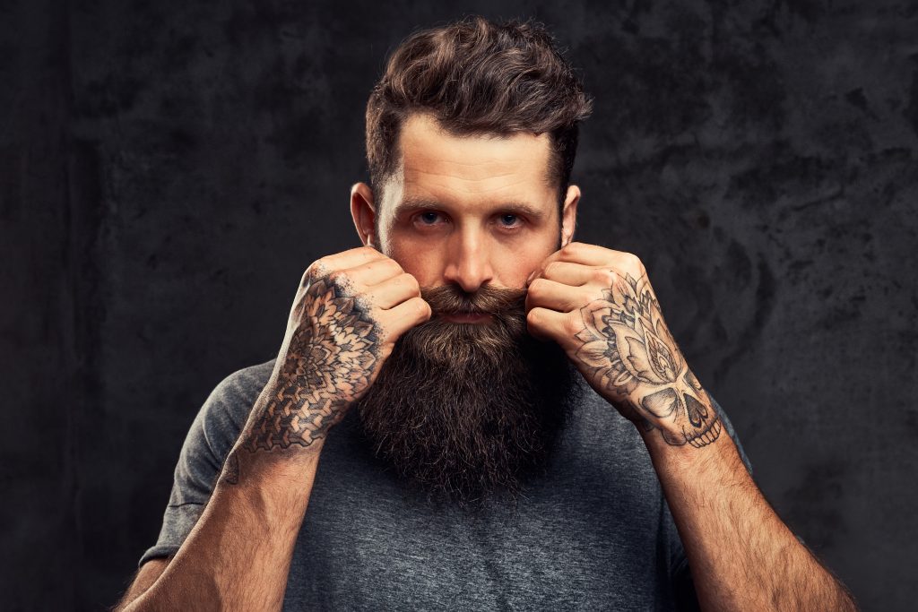 retrato de um hipster tatuado com barba cheia e corte de cabelo elegante vestido com uma camiseta cinza olhando para a camera fica em um estudio em um fundo escuro